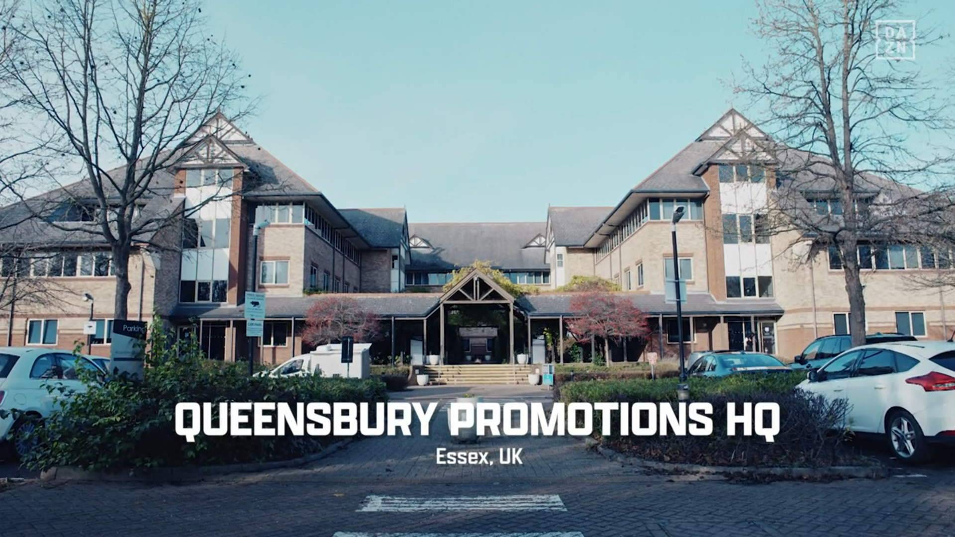 a dull looking building with a dull looking car park on a dull british winter morning, on-screen caption: "QUEENSBERRY PROMOTIONS HQ. Essex, UK"