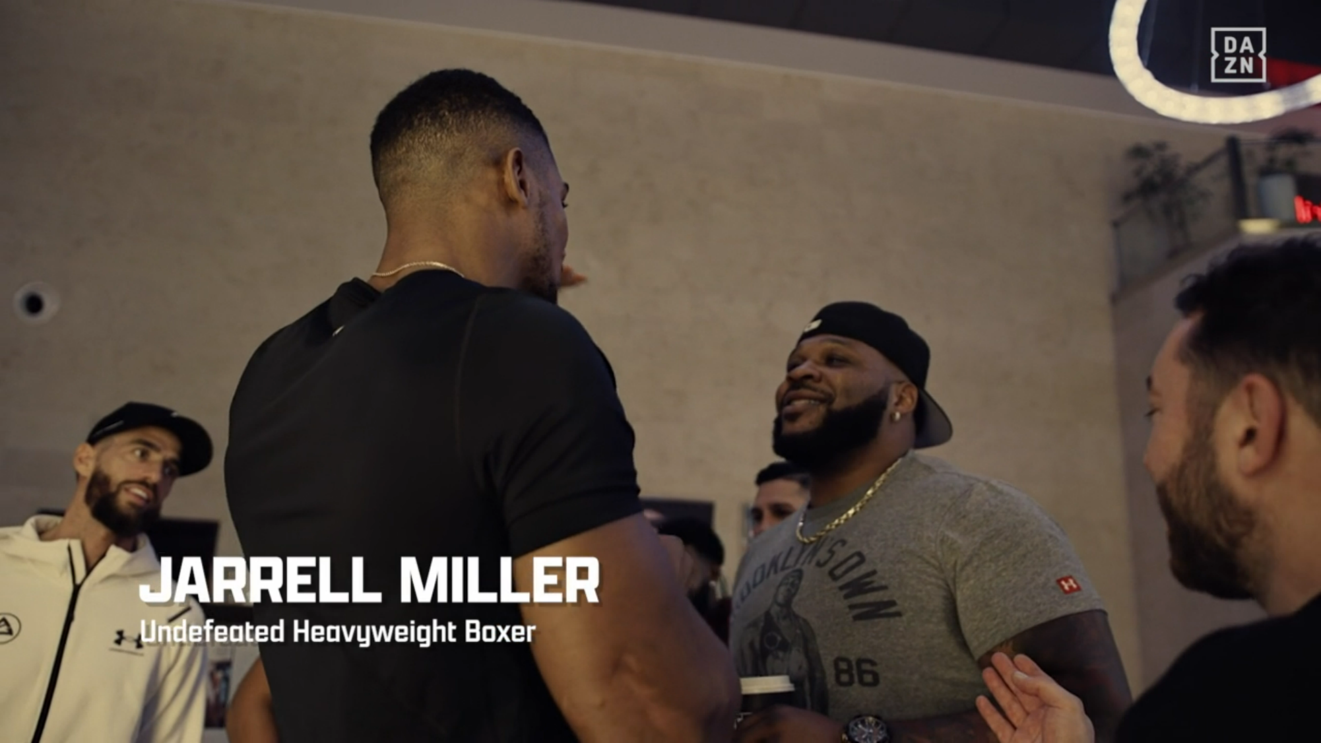 Anthony Joshua meets Jarrell Miller in Saudi Arabia, a man in white in the background has an incredulous look on his face, the on-screen caption says "JARRELL MILLER Undefeated Heavyweight Boxer"