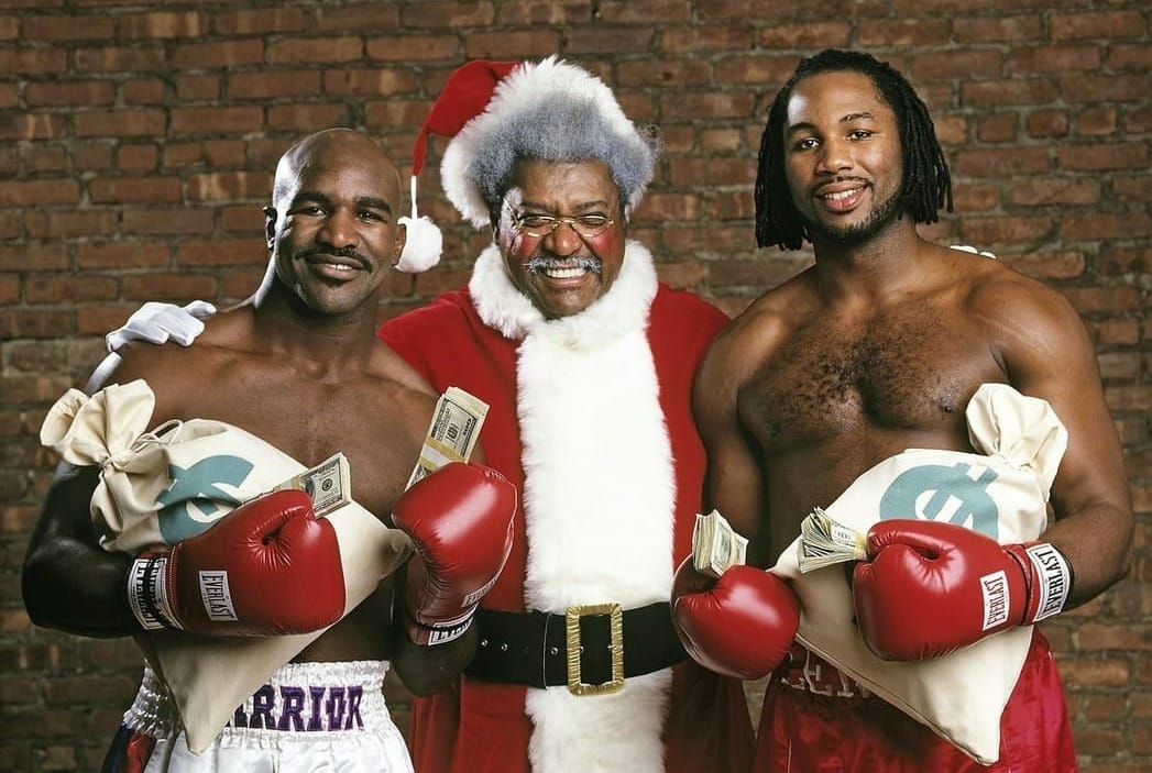 Don King dressed as Santa Claus standing between Lennox Lewis and Evander Holyfield, the two boxers are posing with their christmas presents -- holding sacks of cash and stacks of bills in their arms and boxing gloves