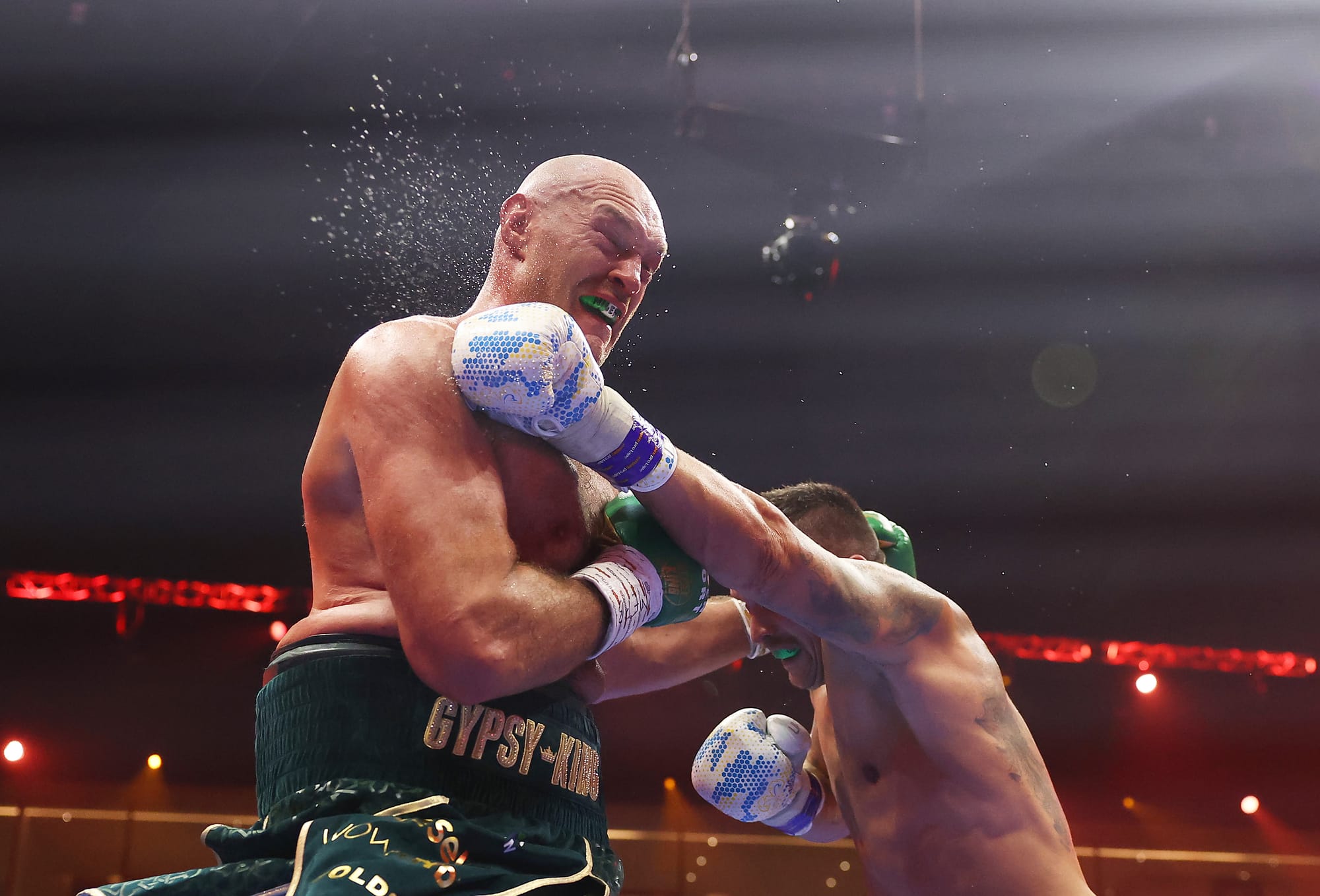 Usyk landing a left straight and sending sweat flying from Fury's head. Fury's expression is pained and his stomach bulges over the side of his boxing trunks.