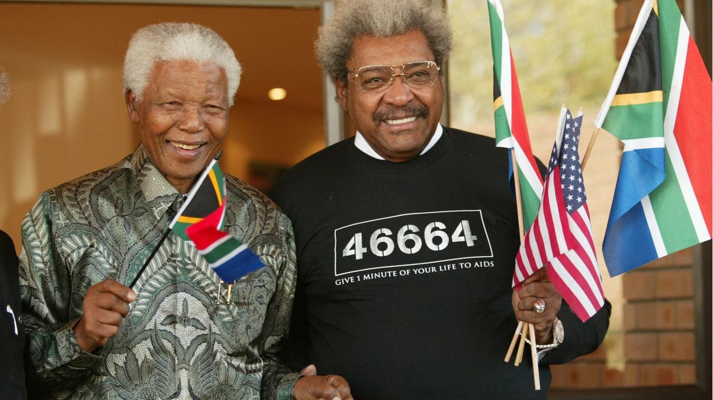 A grey-haired Don King next to white-haired Nelson Mandela. They're waving South African and American flags, King is wearing a shirt in support of an AIDS charity campaign.