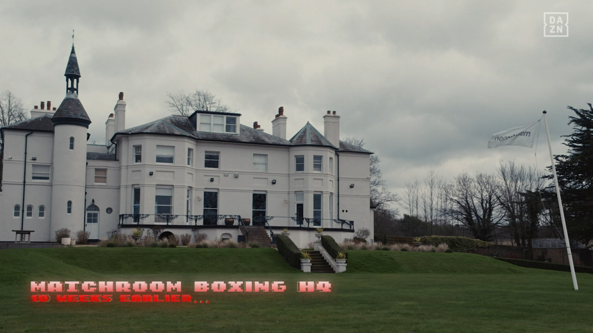 Matchroom Boxing HQ, the building has at least 7 chimneys and there's a flagpole on the front law flying a flying white flag that has "matchroom" written on it in black text. It's a dull, grey, british day.